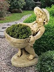 Statue of a young boy holding a large planter filled with green plants, outdoors. The statue is light-colored, and the setting is a garden.