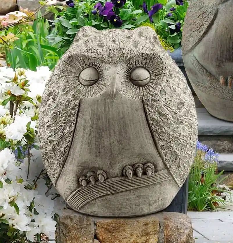 Stone owl statue with closed eyes, perched on a stone ledge amidst a garden with flowers.