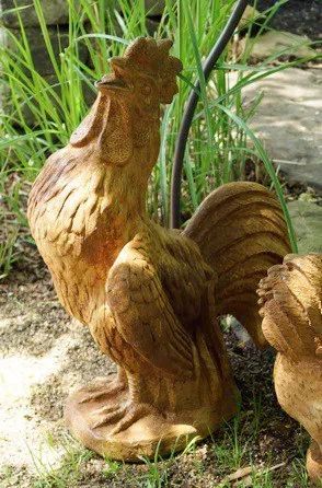 A weathered, brown rooster statue stands outdoors, next to grass and a blurred background.
