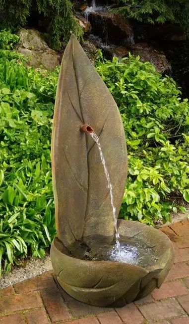Leaf-shaped outdoor water fountain with water flowing from a small spout into a basin, surrounded by greenery.