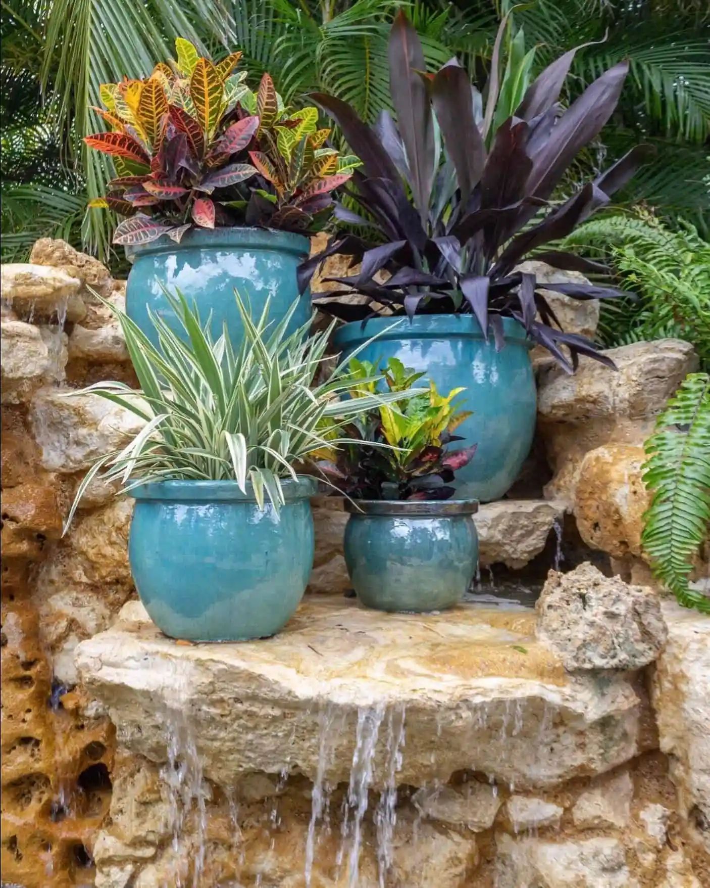 Four turquoise pots with colorful plants sit on a rocky ledge, with water cascading down.