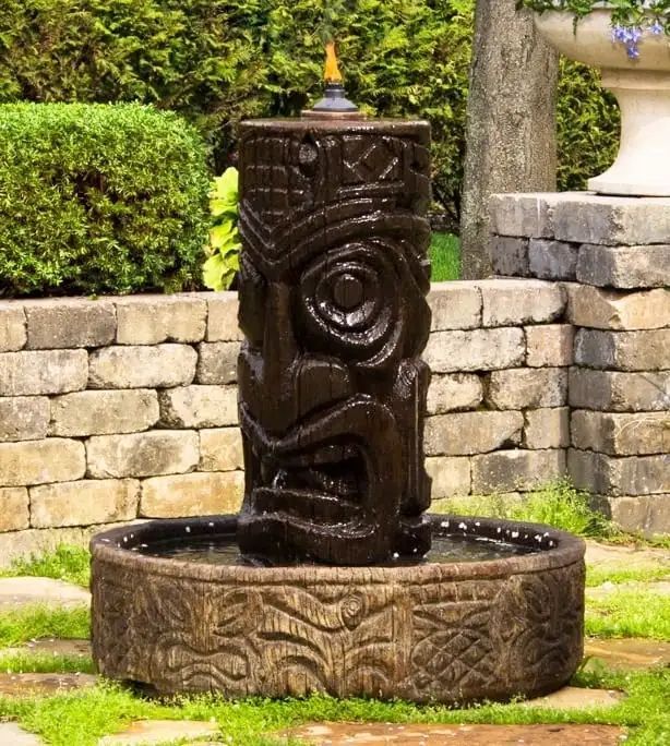 A dark wooden tiki fountain in a stone basin, set outdoors in a garden. The tiki has a carved face and is surrounded by green foliage.