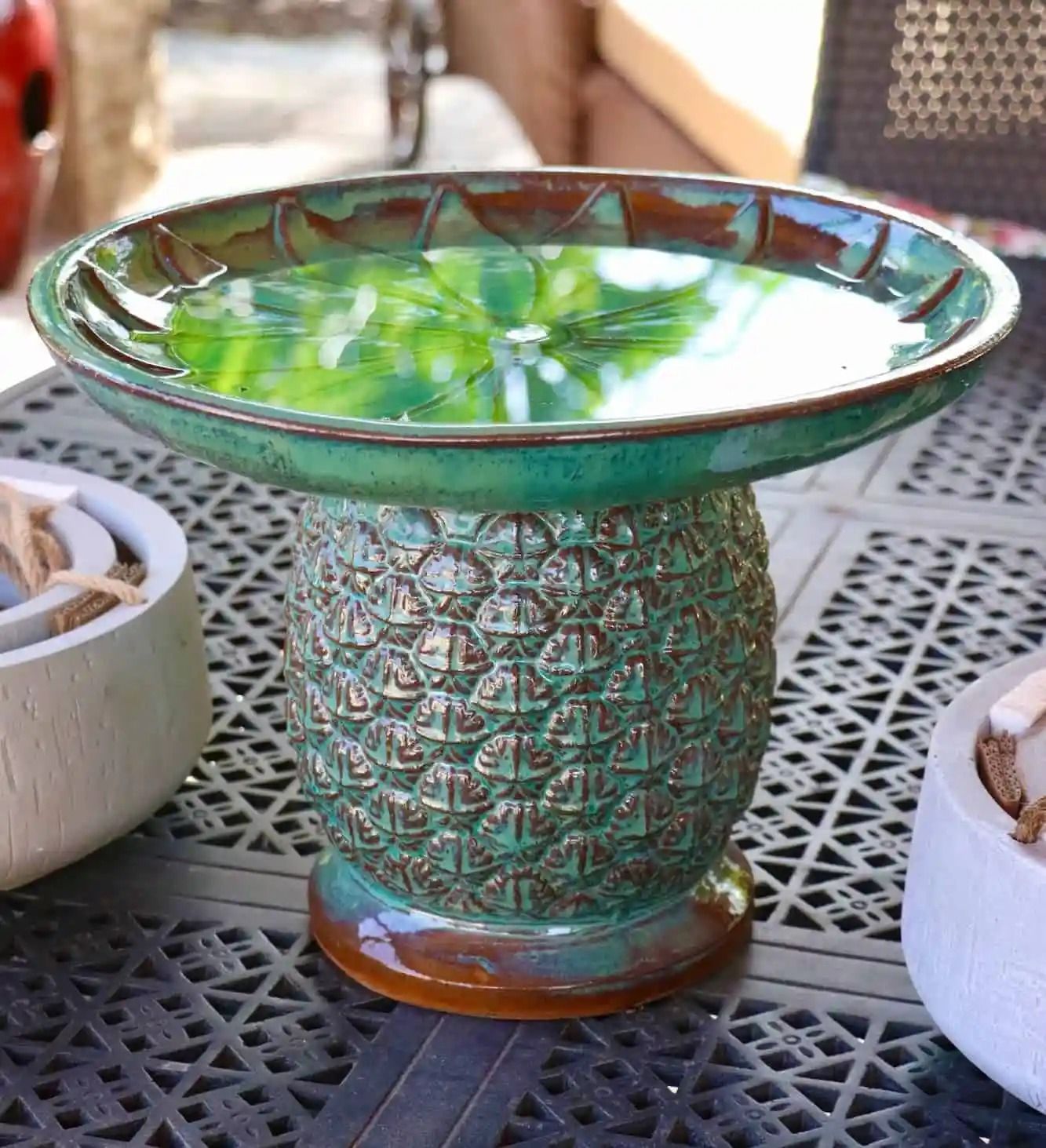 Green ceramic birdbath on a table, shaped like a pineapple. The bowl holds water, reflecting the surrounding greenery.