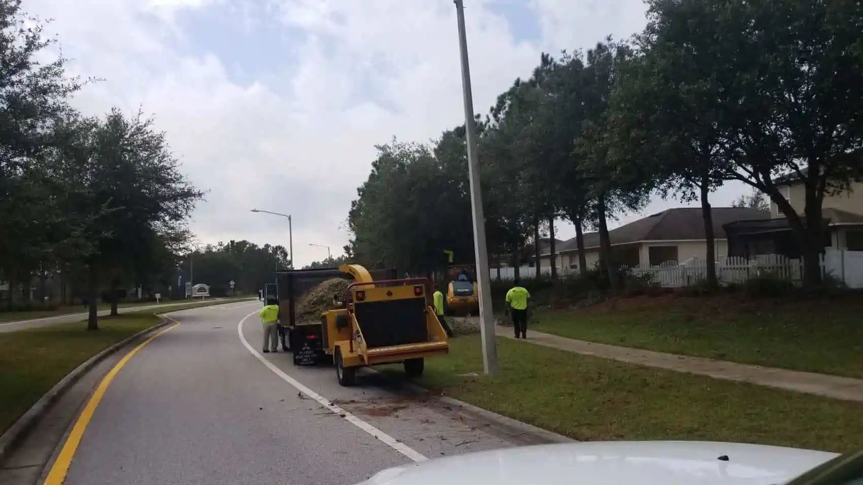 Road maintenance crew using a wood chipper on a street, processing branches. Workers in safety vests, houses in background.