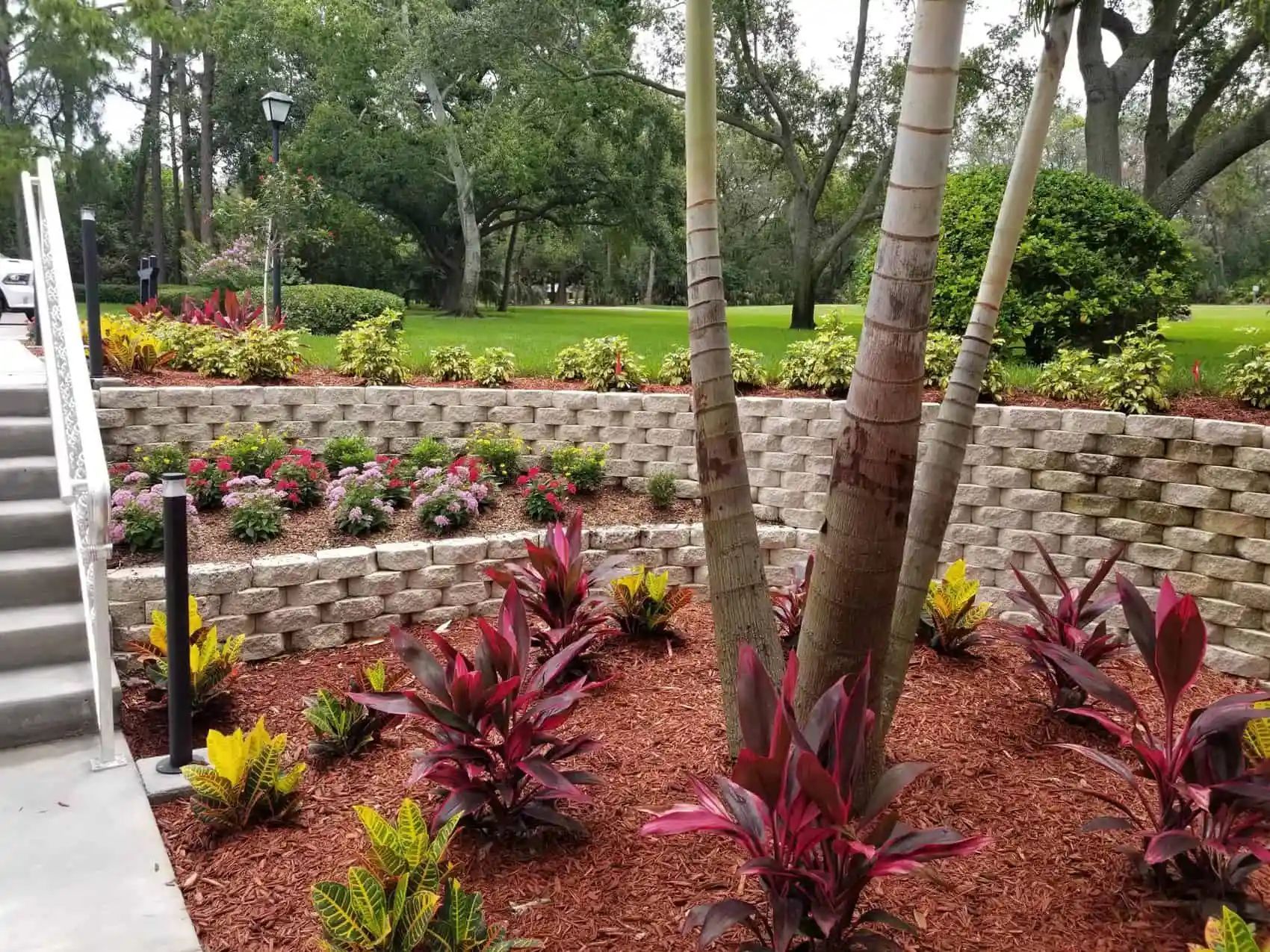 A landscaped tiered garden with red and yellow plants, brick retaining walls, and a stairway. Trees and green grass are in the background.