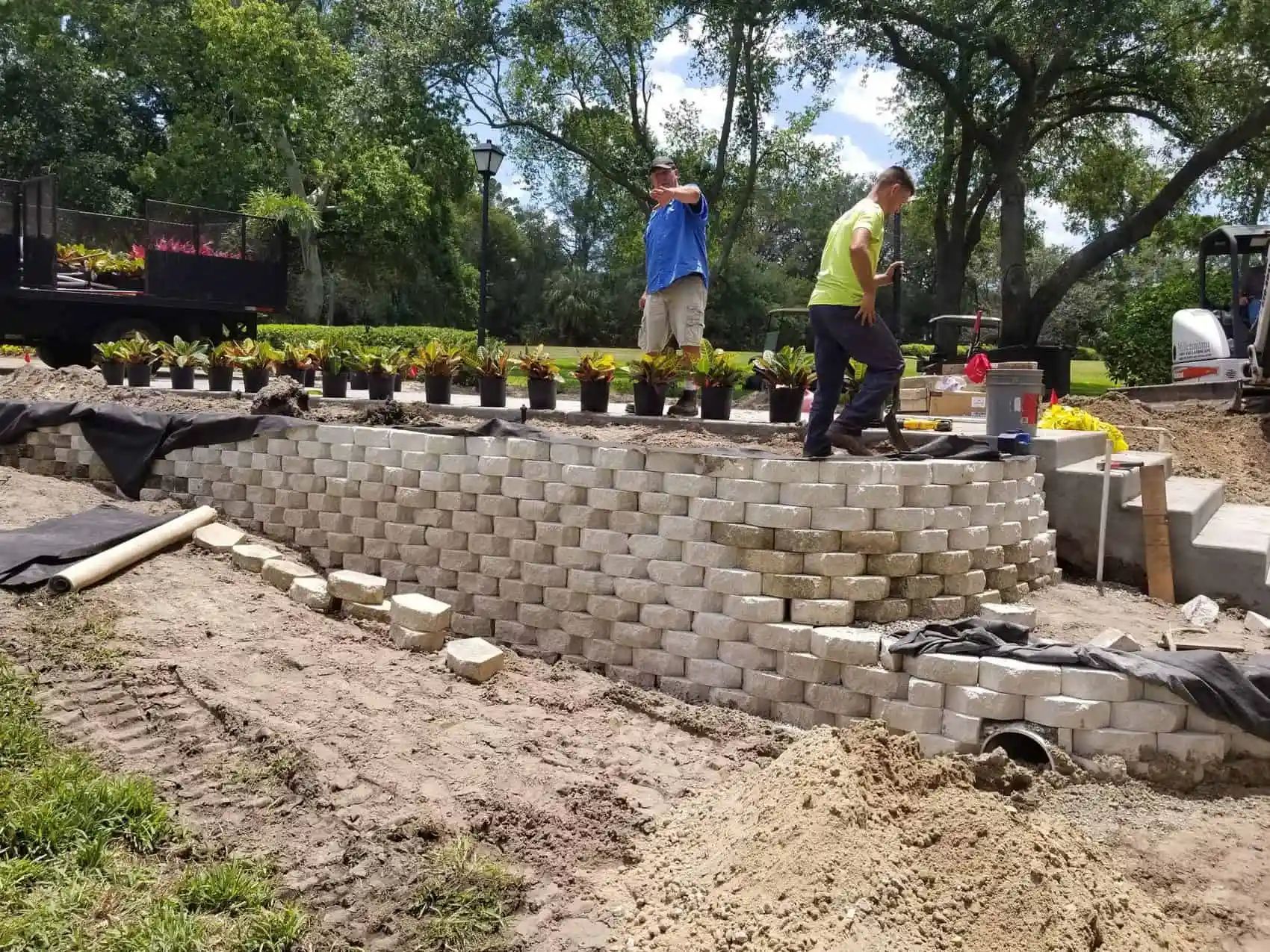 Two men building a retaining wall with concrete blocks in a garden setting. Plants in pots are lined up above the wall.