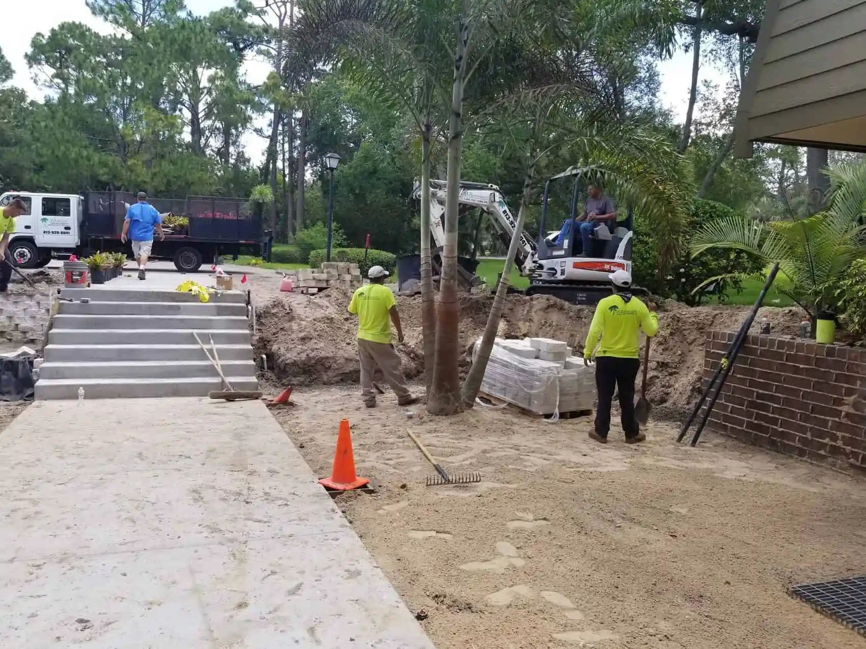 Construction workers building a brick patio and steps. A backhoe, truck, and workers in neon shirts are present in a sunny outdoor setting.
