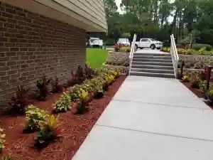 A brick building with a concrete walkway leading to steps. Landscaping with red mulch and plants on either side.