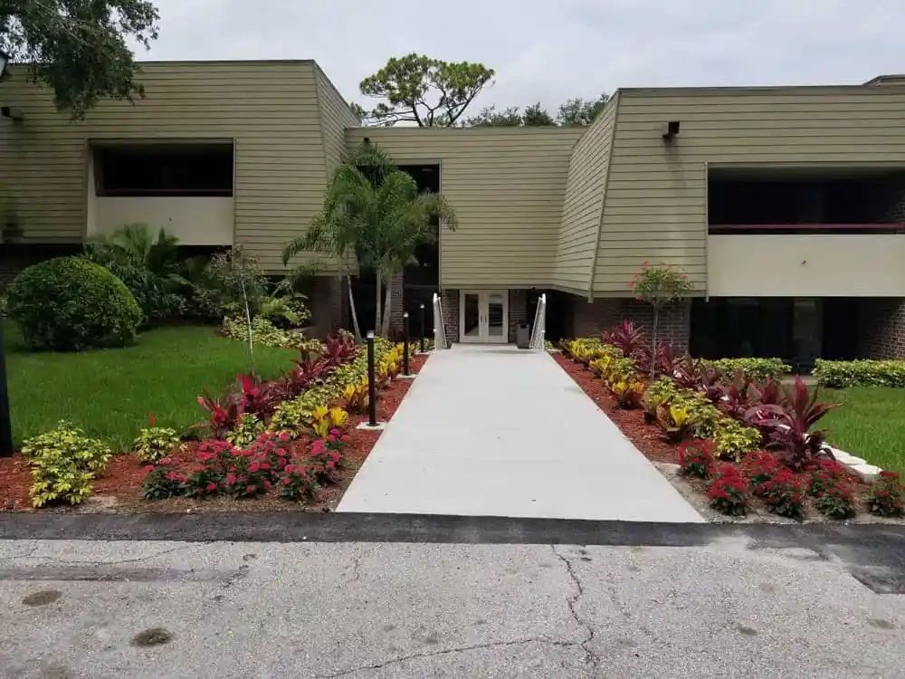 A concrete walkway leads to the entrance of a two-story beige building, flanked by colorful flowerbeds and landscaping.