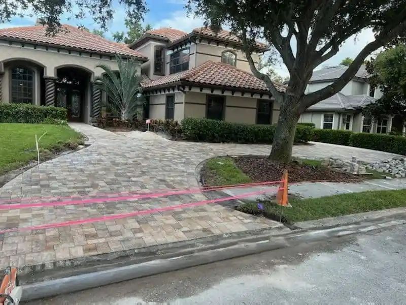 A brick paver driveway in front of a two-story house with a large tree and manicured landscaping.