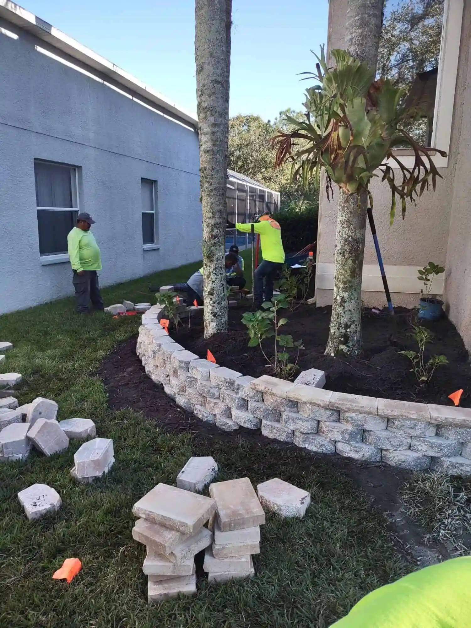 Landscapers building a retaining wall around a garden bed. They are working near a house and palm trees on a sunny day.