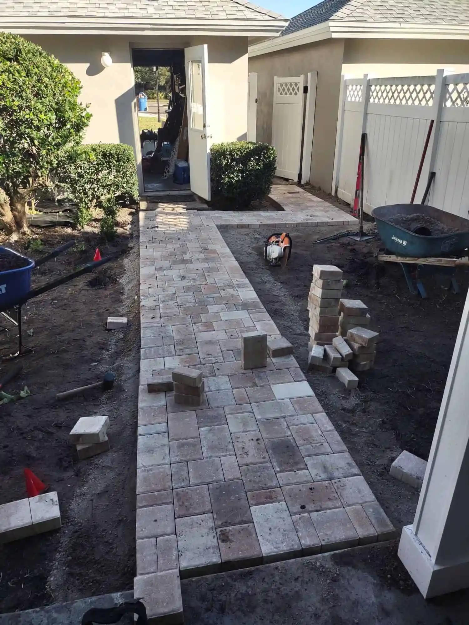 A brick walkway being constructed in a yard, leading to a doorway. The path is partially laid with bricks, surrounded by dirt and tools.