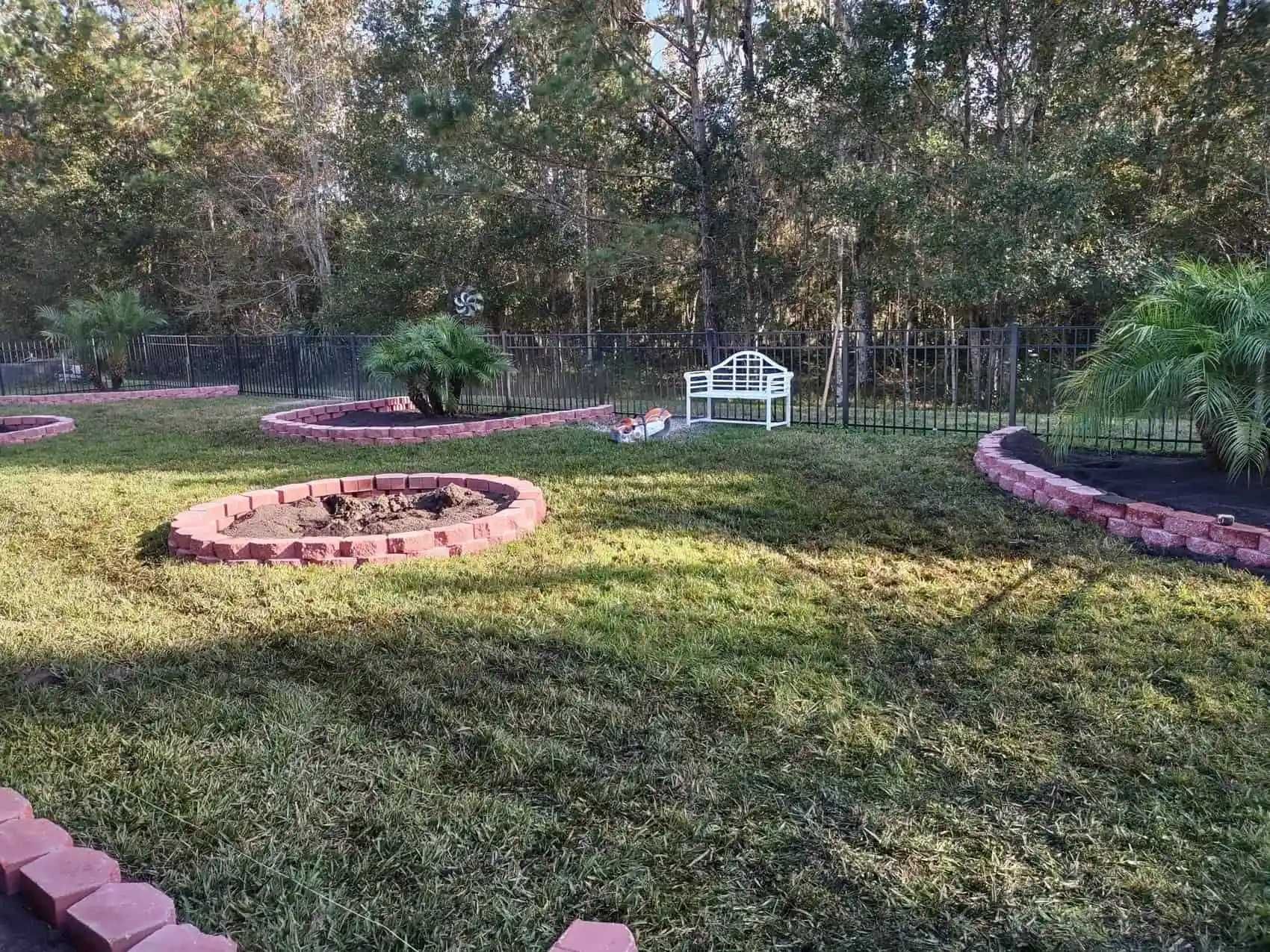 Lush backyard with a brick-lined fire pit, small trees, and a white bench. Dark green grass is surrounded by a black fence and trees.