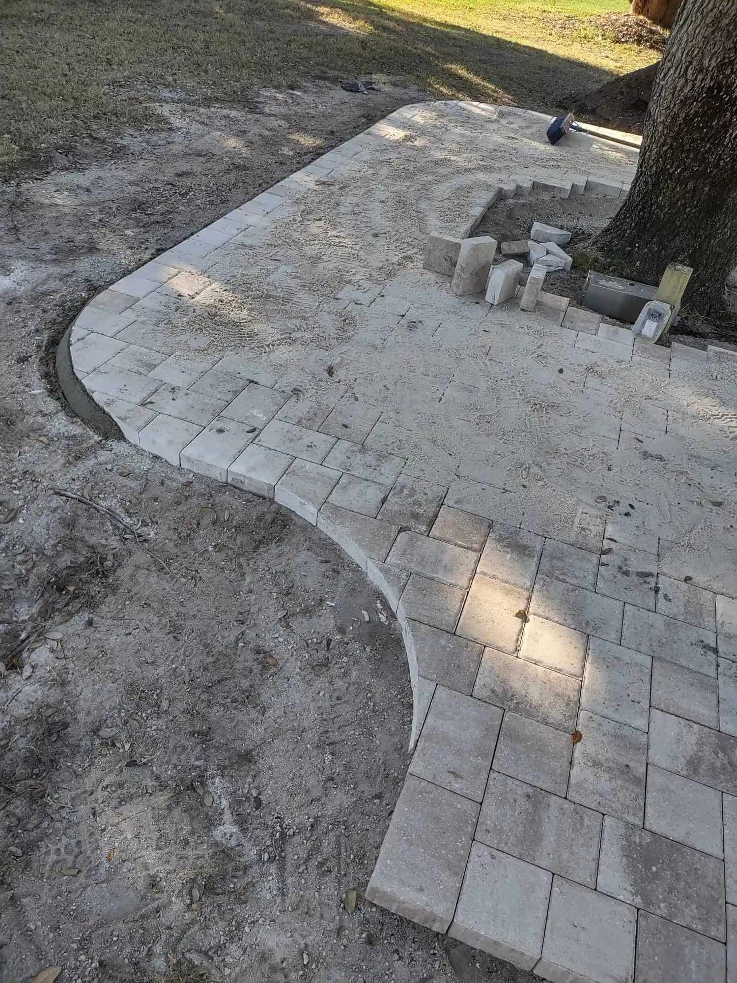 A partially-built brick patio curving around a tree trunk, set in a dirt yard. Some bricks are stacked nearby.