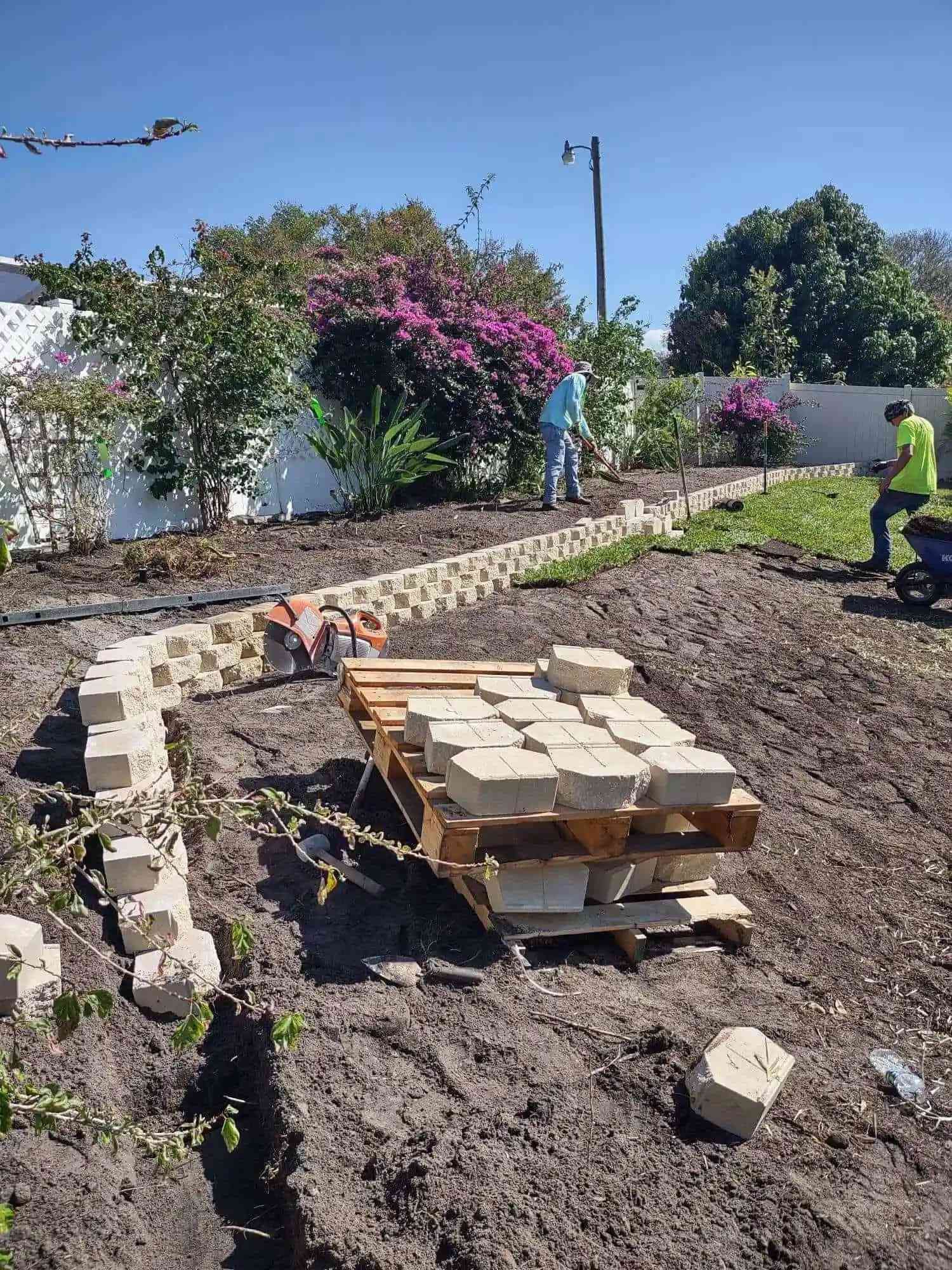 Construction of a retaining wall with beige blocks on a hillside. Two workers and a pallet of blocks are visible.