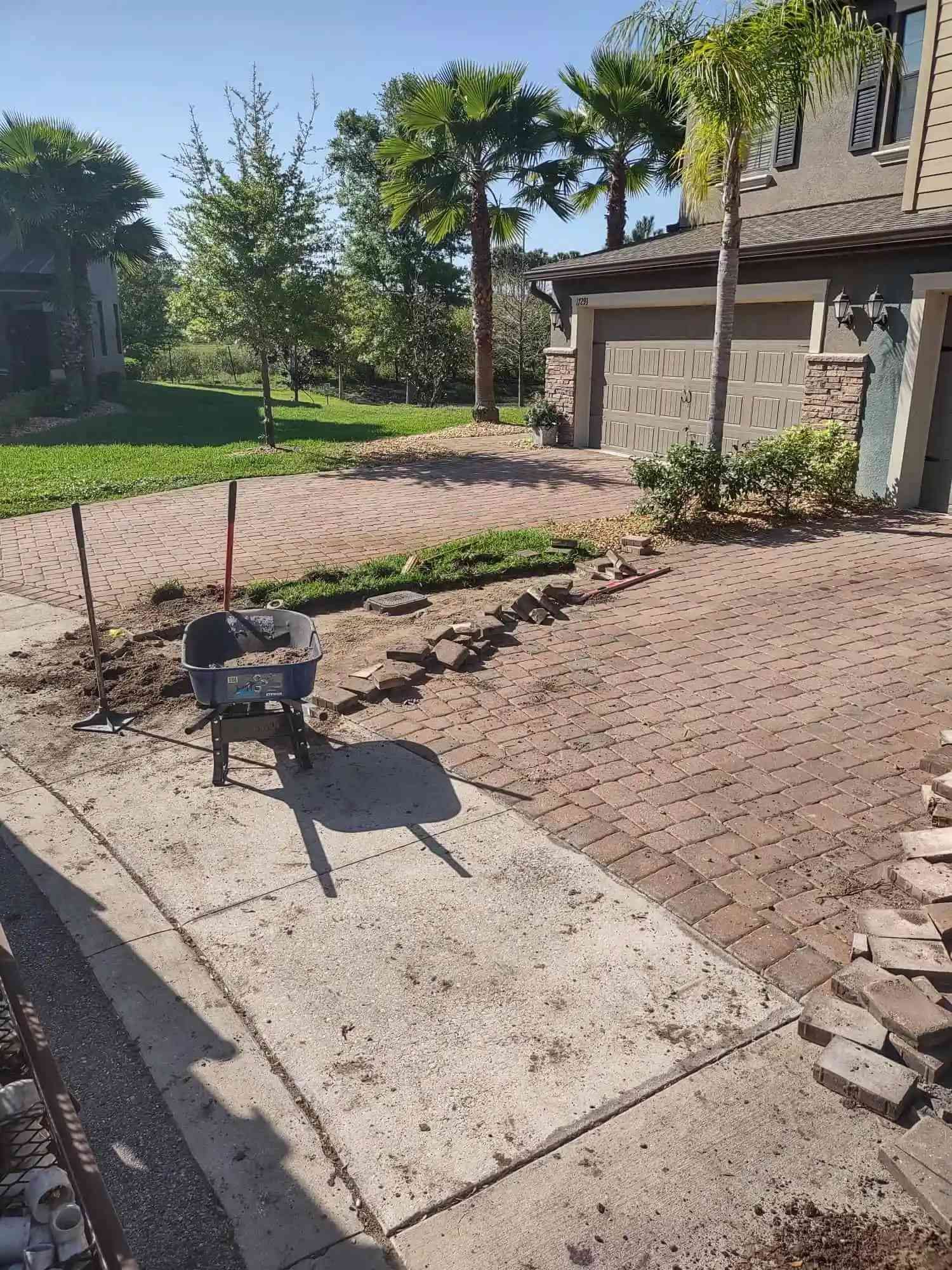 A brick driveway under construction with a wheelbarrow, tools, and loose bricks. The setting is near a house and street, with trees in the background.