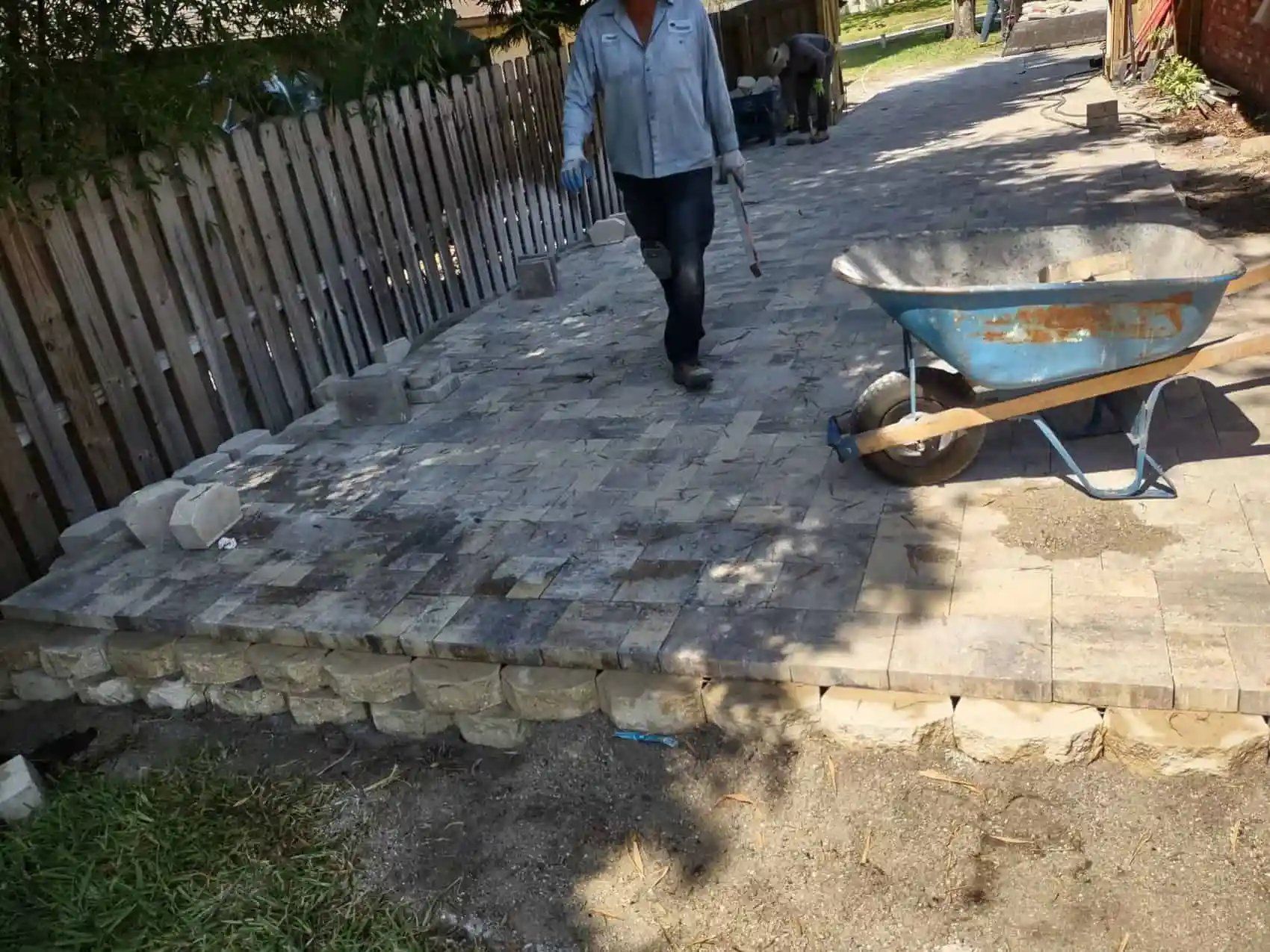 Person walking on newly paved walkway with retaining wall and wheelbarrow. Wooden fence on the left.