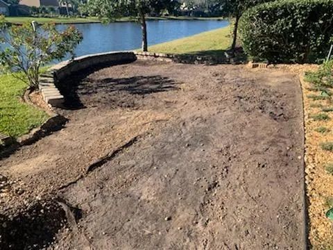A dirt area with a curved stone wall and view of a lake. There's green grass and some small plants.