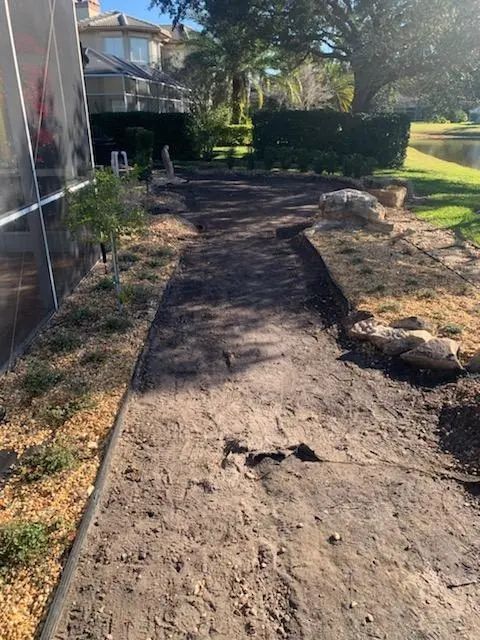 A dirt path in a backyard bordered by a screen enclosure, landscaping, and a grassy area. The path appears prepared for planting.