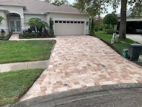 Brick driveway leads to a beige garage door of a suburban home with a well-manicured lawn.