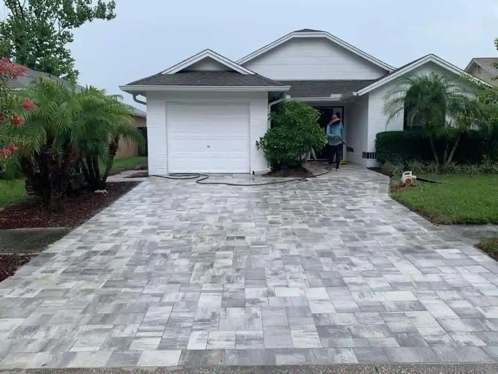 A house with a gray brick driveway. A person is visible in the doorway. Palm trees and shrubbery decorate the front yard.