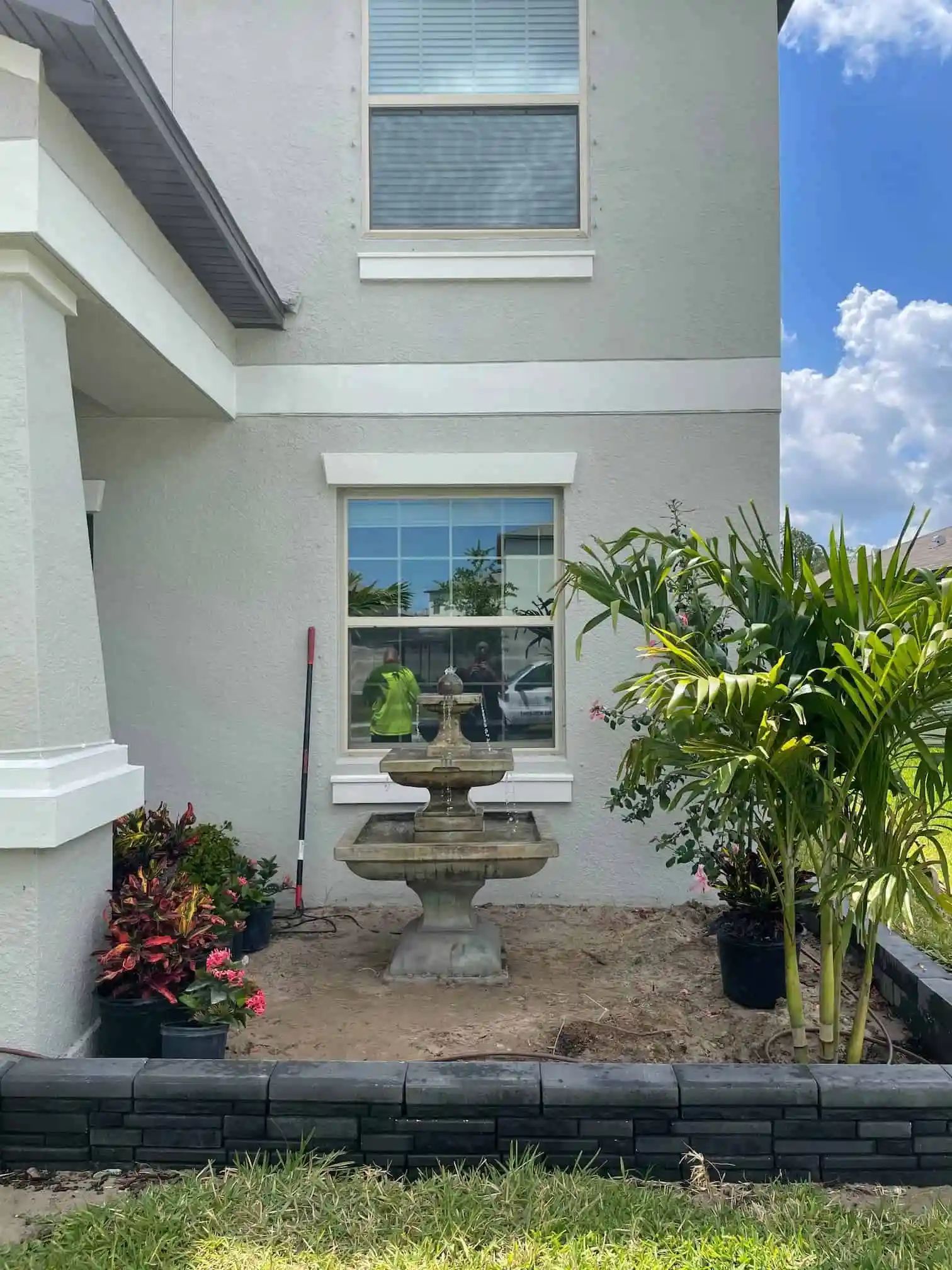 A gray stone fountain sits in a bare flowerbed in front of a light gray house. A window is centered behind the fountain.