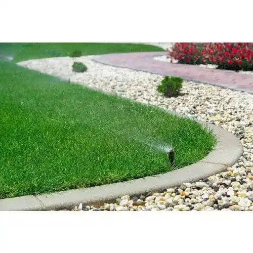 Lawn sprinkler watering a green grass lawn next to a rock border and a paved path.