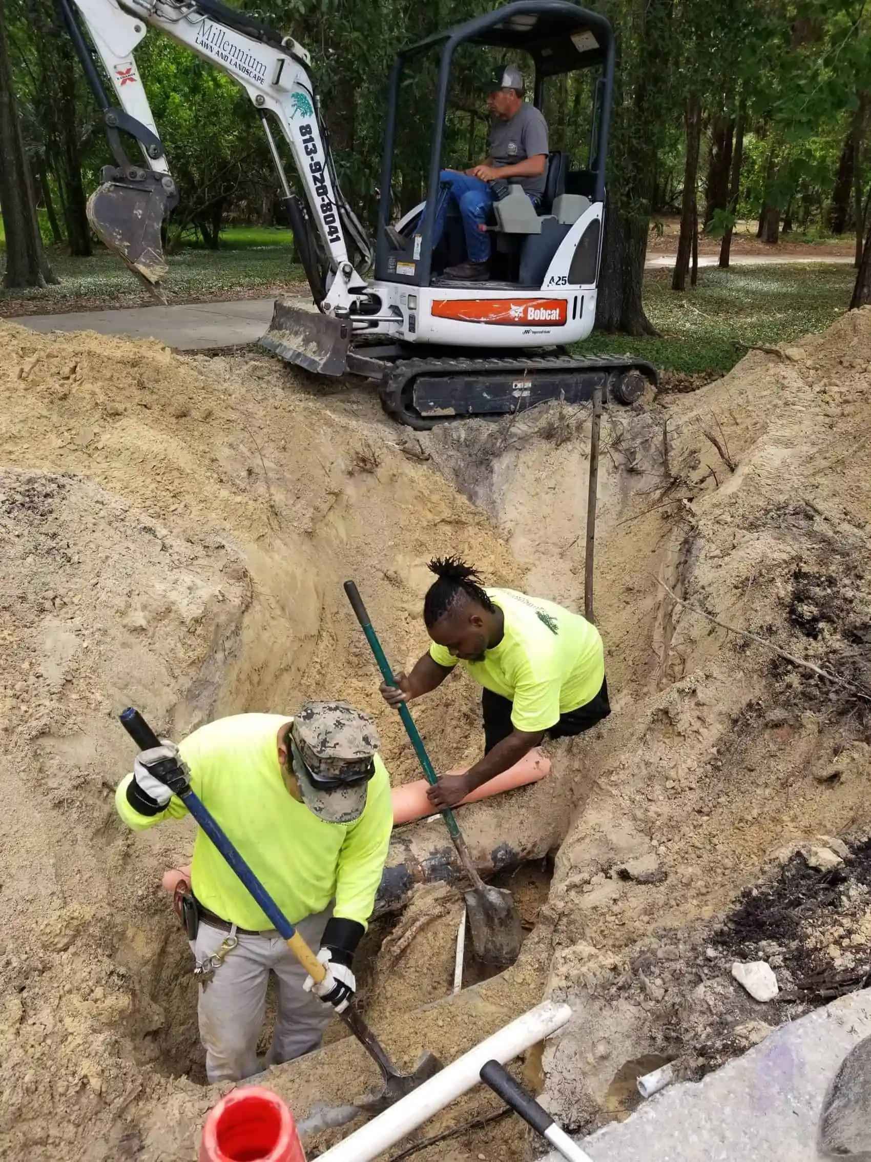 Construction workers digging a trench with shovels, while a Bobcat excavator sits above. The workers wear neon yellow shirts and are outside.