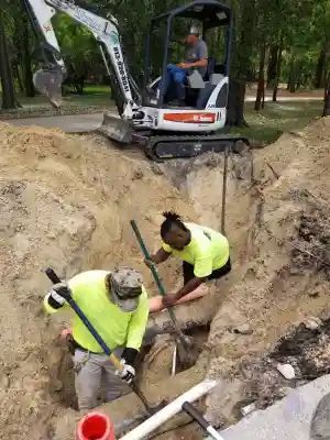 Construction workers use shovels to work in a deep trench, as a small excavator sits above them. They are wearing neon yellow shirts.