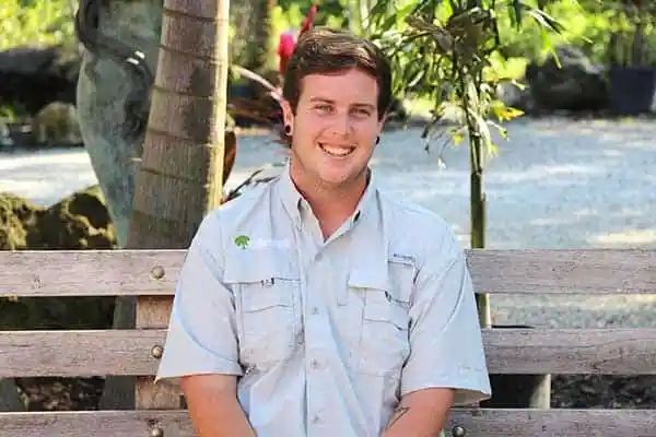 Man with short brown hair, smiling, in a light-colored shirt, sitting on a wooden bench outdoors.