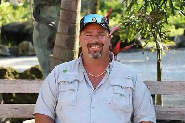 Man with a beard and baseball cap smiling outdoors, wearing a light-colored shirt and gold chain, sitting in front of trees.