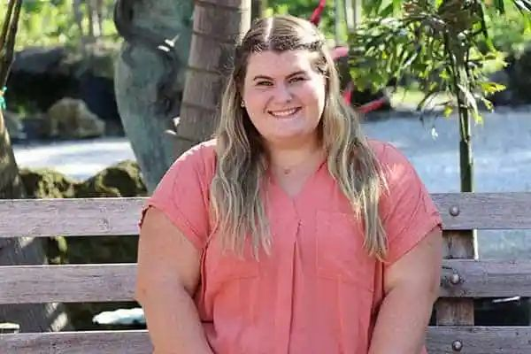 Woman with blonde hair smiles while sitting on a wooden bench in a garden setting. She wears a coral-colored top.