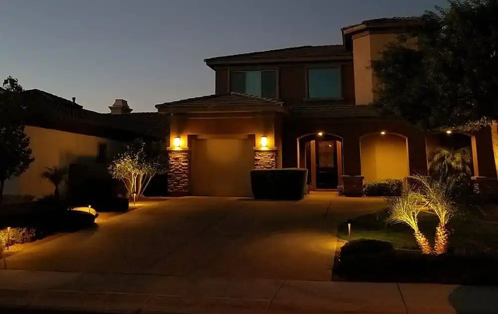 A two-story home at dusk, lit with warm exterior lights illuminating the facade, driveway, and landscaping.