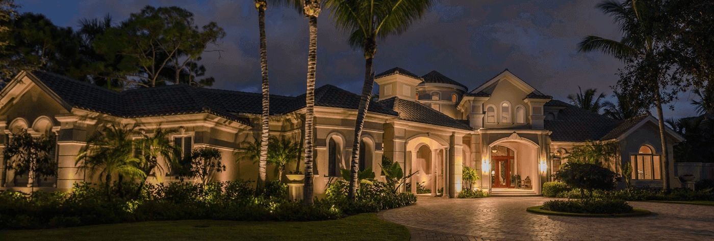 Large, illuminated mansion at dusk, with palm trees and landscaping. The house features multiple stories, ornate details, and a circular driveway.