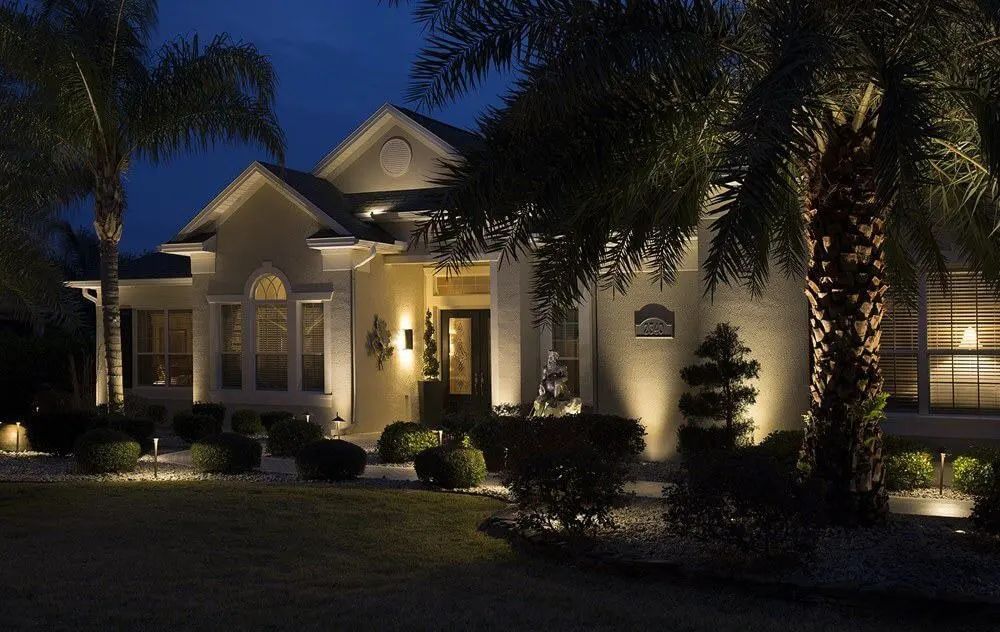 A well-lit house at night with spotlights on landscaping and palm trees.