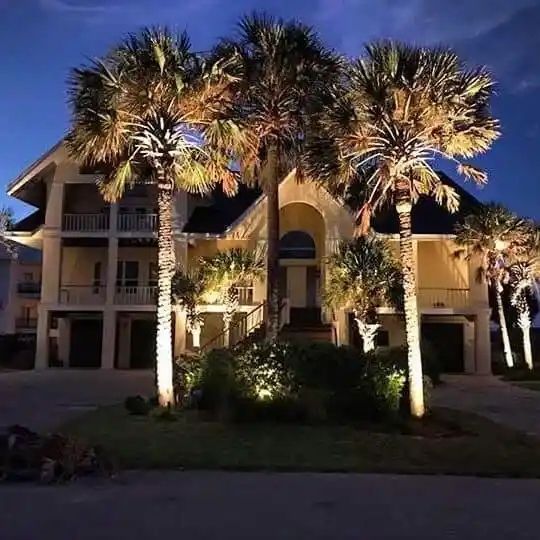 A multi-story beach house at night, illuminated with spotlights on palm trees and shrubbery.