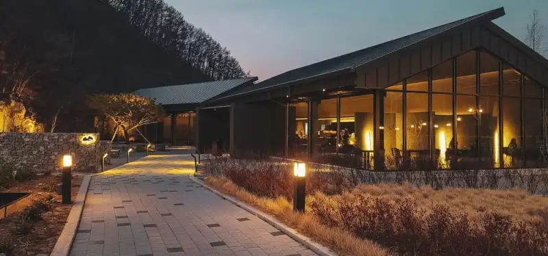 A modern building with large windows is lit up at dusk, with a pathway leading to it and a mountain in the background.