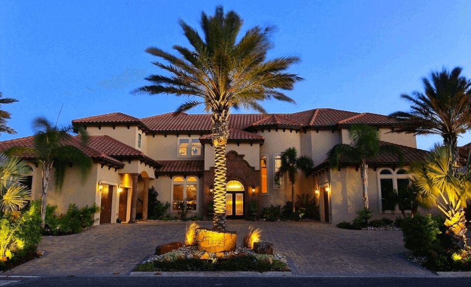 Luxury home at dusk with palm trees, lit landscaping, and a brick driveway. The house has a red tile roof and arched windows.