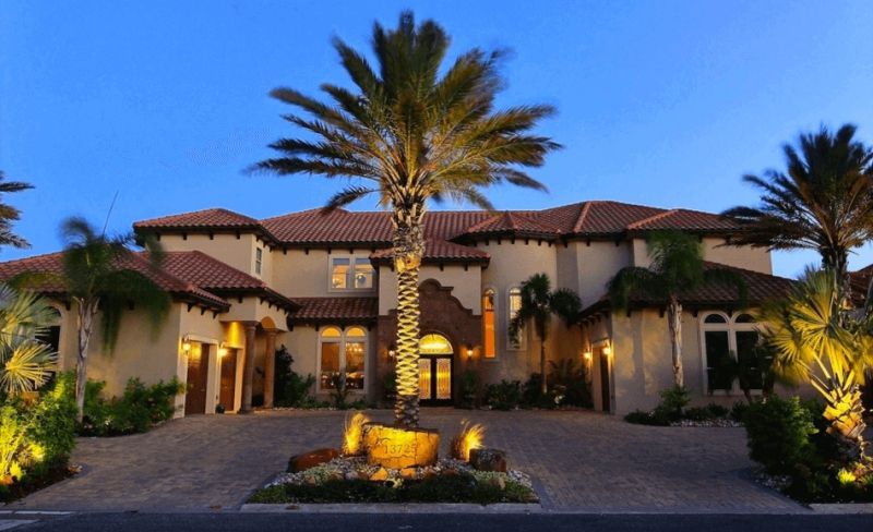 Large, luxurious two-story home with a palm tree in the center of the driveway, lit up at dusk. Beige walls, red-tiled roof, and decorative lighting.
