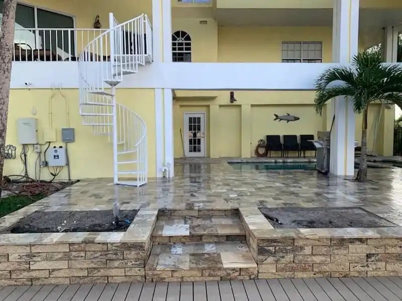 Exterior view of a light yellow house with a white spiral staircase, a pool area, and a stone retaining wall. The area is paved in stone.