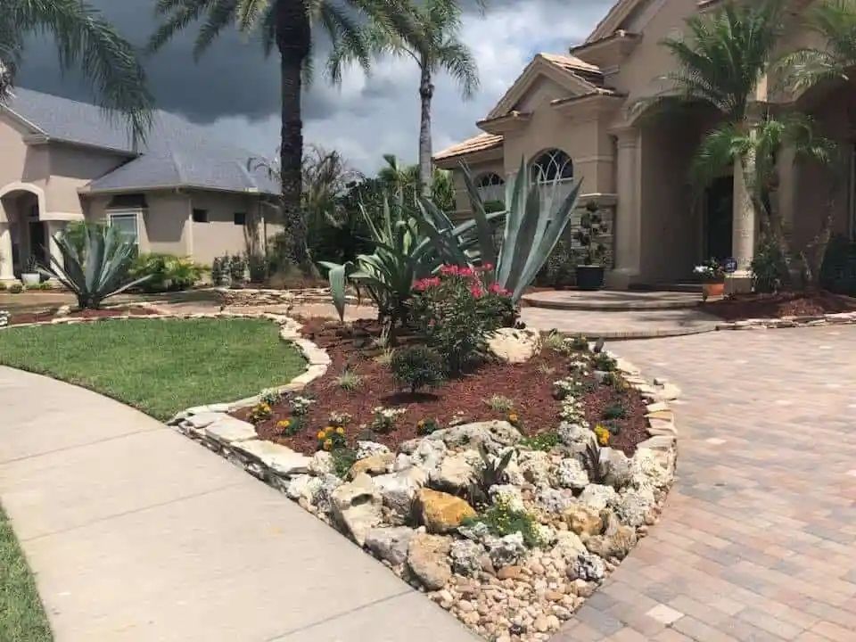 A landscaped front yard with a variety of plants, including agave and flowers, bordered by stone and mulch, with a sidewalk in the foreground.