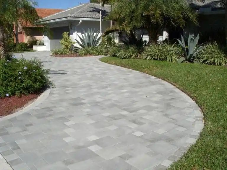A paved driveway curves towards a white house with a manicured lawn and landscaping. The driveway is gray, the lawn green.