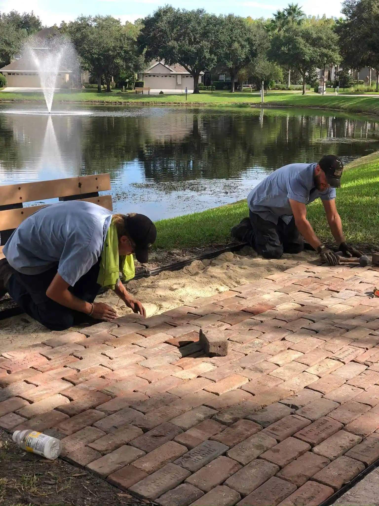 Two workers laying bricks on a pathway near a pond with a fountain. One is kneeling and the other is bent over.