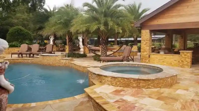 A backyard with a pool and hot tub, palm trees, and a wood-paneled structure. Lounging chairs are scattered around.
