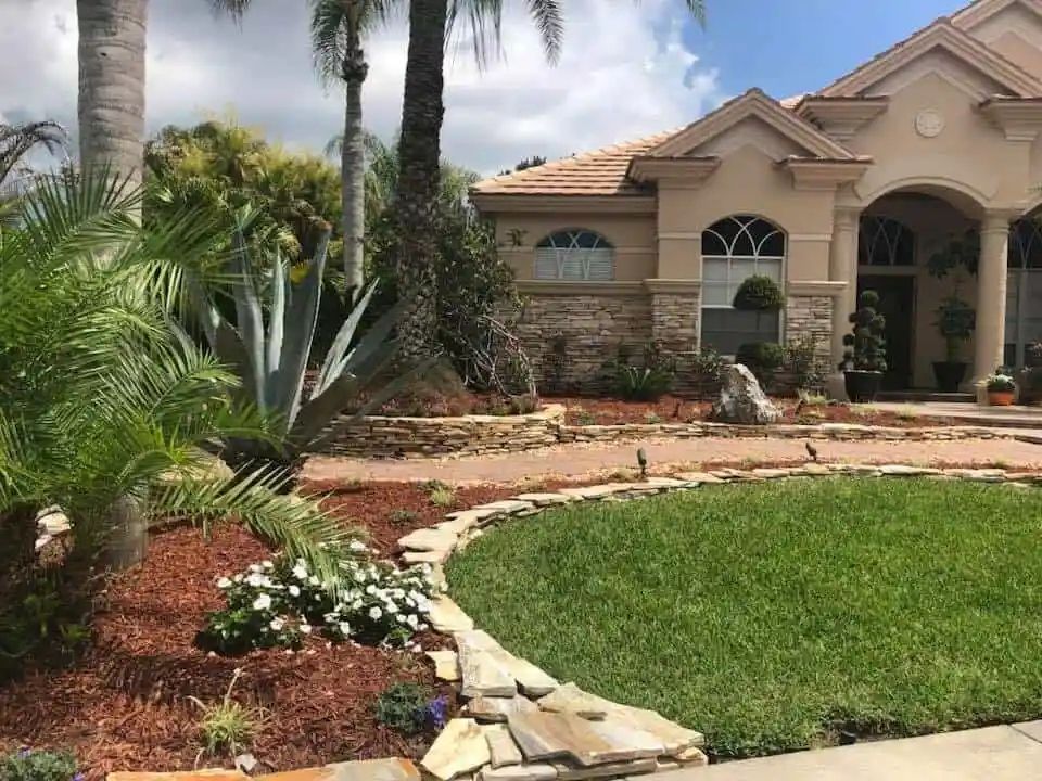 A well-manicured home with a stone facade, lush green lawn, and tropical landscaping. The scene is sunny, with blue skies peeking through.