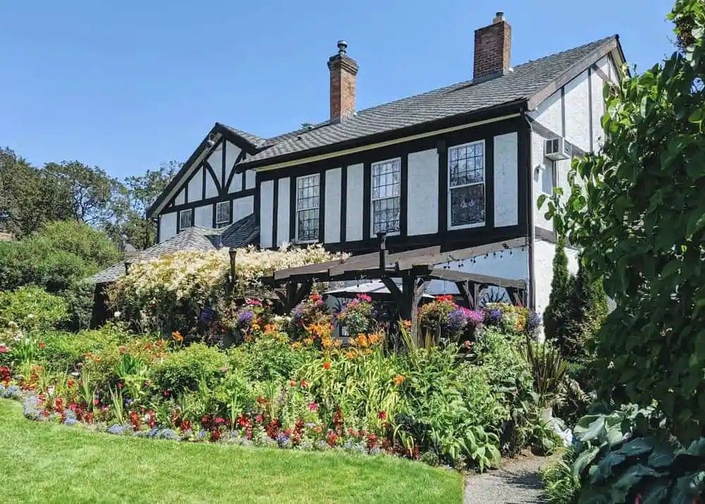 A two-story Tudor-style building with black and white facade, surrounded by a colorful flower garden and green lawn under a blue sky.