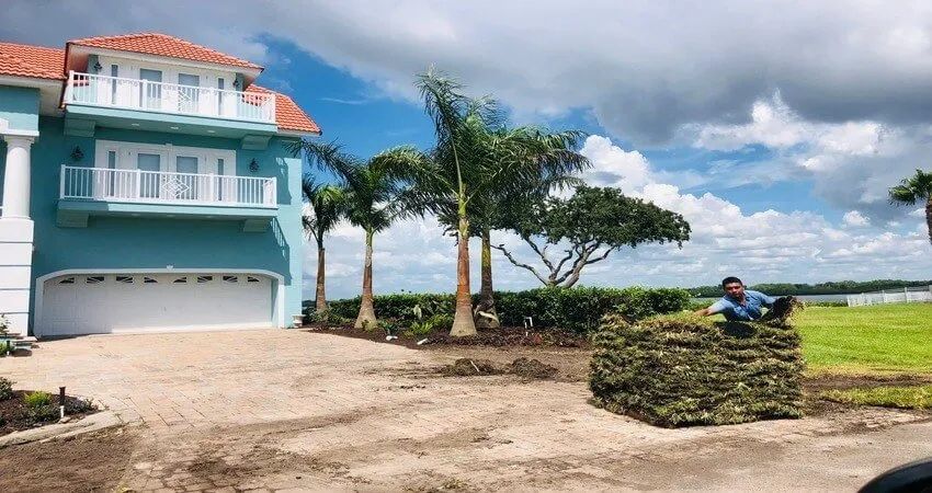 A person stacking sod in front of a teal house with a white garage, palm trees, and a lake under a cloudy sky.