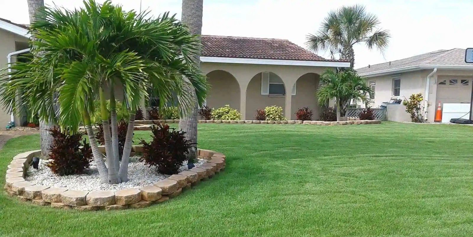 A house with a lush green lawn, a bed of white stones and dark red plants surrounding a palm tree.