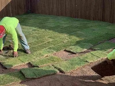 Two workers laying rectangular grass sod in a backyard. The workers wear neon green shirts and the sod is green.