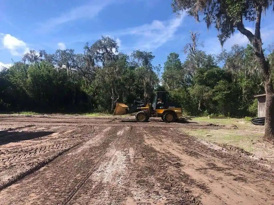 A yellow loader on a muddy lot, clearing a space near a wooded area under a blue sky.
